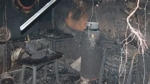 A burnt and blackened scene, showing the remains of a gas canister and heater