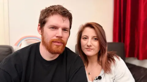 A picture of Corrinne and Sam Boulton sat on a sofa. Sam has a ginger beard and brown hair and Corrinne has shoulder length brown hair.