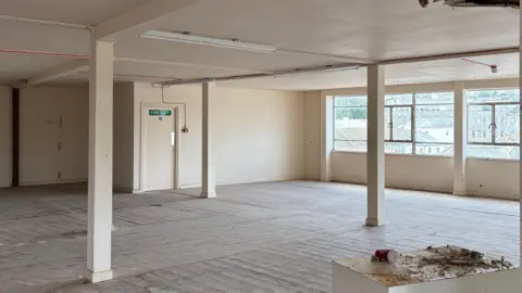 An empty derelict white room with ground-to-ceiling pillars and with large windows in the background.