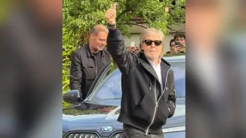 Paul McCartney waves to the crowd. He is smiling and wearing sunglasses. He is wearing a light coloured top, black jeans and a black hooded jacket. He is standing in front of a car and some people can be seen in the background, including someone filming on their mobile phone.