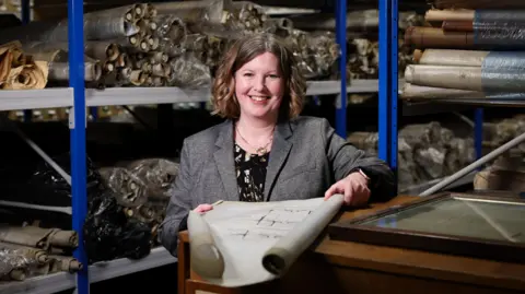 A woman with shoulder-length curly brown hair smiles at the camera. She is standing in what looks like a store room holding a large piece of paper, which looks like a blueprint. She is wearing a grey blazer. 