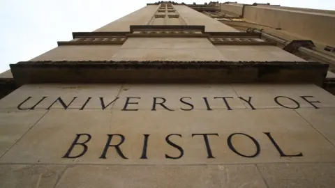 A shot pointing up the height of a University of Bristol building. 'The University of Bristol' can be seen spelled out in the beige stone of the building's wall which reaches up to the sky.
