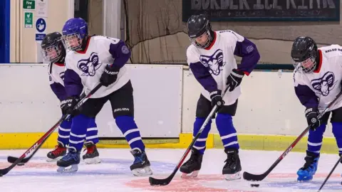 Kelso Rams Ice Hockey Club Four young ice hockey players in a line on ice skates, wearing blue helmets, white ice hockey shirts with ram head in centre, holding hockey sticks out in front.