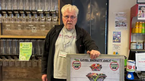 LDRS A man with white hair and glasses is stood in a shop with empty bottles behind him. A sign in front of him instructs people to have level baskets only.