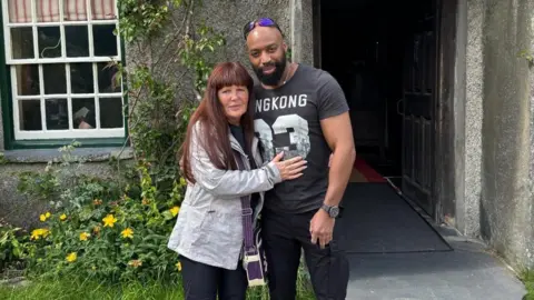 Denise, with red hair and a light blue jacket, with her arms around her partner Sylvester Abayomi, wearing a black T-Shirt with white numbers and with sunglasses on his head, in a photo released by the family.