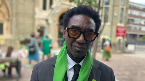 Damel Carayol, from Humanity for Grenfell, wearing a charcoal suit, green scarf and has tinted glasses and a short afro.
