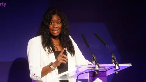 PA Media The minister for Devolution, Faith and Communities Miatta Fahnbulleh is wearing a white jacket and delivering a speech in front of a glass lectern with a purple backdrop.