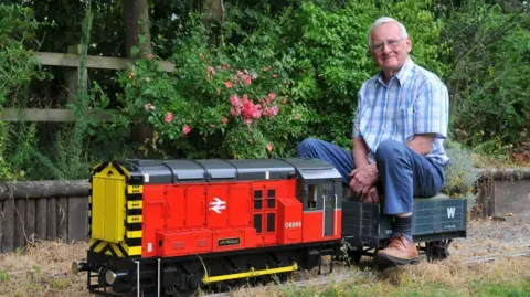 A smiling Richard Periam is seen sitting on the rear carriage of a model train. He is wearing a checked shirt and blue trousers. The rest of the railway carriage is red with a yellow radiator