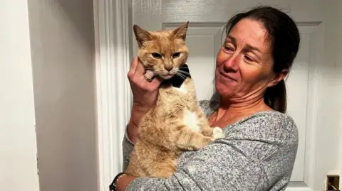 Lorraine Clarke, who has brown hair tied back and is wearing a grey sweater, holding a ginger and white cat. She is standing inside a home, in front of a white door. She is looking at the cat and stroking him.
