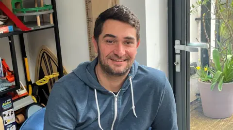 George Kokkinos looking at the camera and smiling while sat indoors. There is a large glass door in the background. He is wearing a blue hoodie.