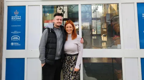 Good Shepherd Mitch at Central Community Shop with Service Manager at Good Shepherd, Chloe Bladen. A man and woman standing together outside a shop. They are positioned in the centre of the frame, standing close, with one person’s arm around the other. Behind them is the entrance to a building with a large blue sign that reads:“Central Community Shop”. The individual on the left is wearing dark trousers, a padded sleeveless jacket over a hoodie, and trainers. The individual on the right is wearing a long patterned skirt and a light long‑sleeved top. They are standing on a paved area directly in front of the doorway.