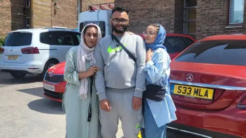 Handout Jamil Hussain standing with his mum and sister and posing for a photo. He is wearing a full grey Nike tracksuit and a black cross body bag. His mother is wearing a light blue abaya dress with a white headscarf. His sister has braided hair and is wearing blue jeans, a blue oversized shirt and a blue headscarf. They are standing in a car park on a sunny day.