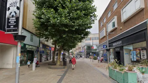 General view of Dudley Street in Wolverhampton. It shows two rows of shops on opposite sides of a paved area. A Holland and Barrett shop is visible on the left and a Metro bank in the background. There are a handful of people walking around