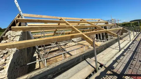 B Russell The roof under construction, with its long wooden beams visible and arranged as part of the roof structure.