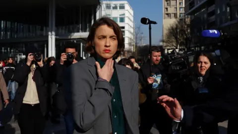 Adèle Haenel wearing a dark grey suit jacket and a dark green shirt underneath. She is standing outdoors, squinting in the sunshine, and is surrounded by journalists taking photos of her