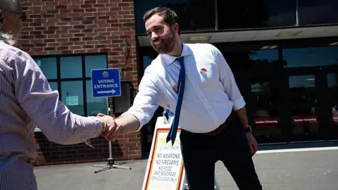 Getty Images Man in white button-down shirt and black pants leans forward to shake a person's hand while standing in front of a building, as his tie swings out.