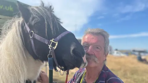 George Carden/BBC Simon who has grey hair and moustache kneeling next to his pony Obama who pulls the cart along. Obama has black and white hair.
