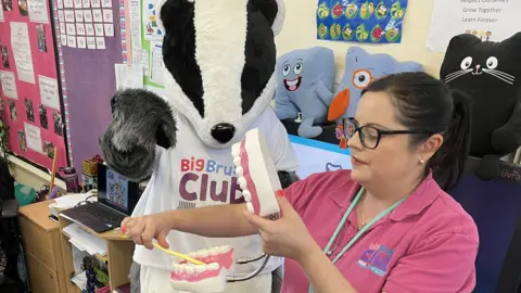 Jermaine Badman from At Home Dental brushing a large model of a set of teeth with a toothbrush standing next to the club mascot - Bob the Badger