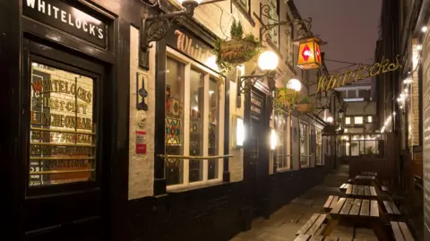 A black and white painted building with several windows, some stained glass. There is a metal sign hanging outside which says 'Whitelocks'. The building is located in an alleyway with wooden benches and tables outside.