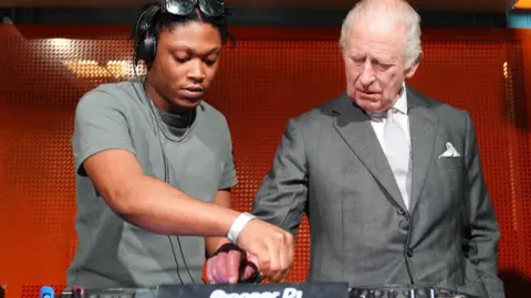 Christian St Louis helps King Charles operate his DJ deck during a visit to Aviva Studios in Manchester, the home of Factory International. Charles is wearing a light grey suit with a white shirt and a pale grey tie.