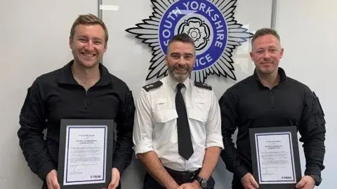 PC Ben Hawley (left) and PC Carl Holden (right) pictured, smiling and holding their awards, with Doncaster District Commander Ch Supt Pete Thorp (Middle) in front of the South Yorkshire Police emblem.