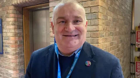 Gary Woodman is standing in a council building, wearing a blue suit. He has short, light hair and wears a blue jacket over a blue jumper.