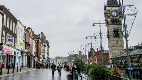 High Row in Darlington town centre, with shops on one side of the pedestrianised street, where people are walking, and an ornate clock tower on the other side. The ground is wet and the sky is cloudy. Unlit Christmas lights are attached to the lampposts. 