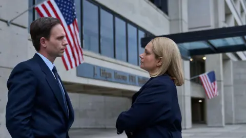 BBC/World Productions Two actors in dark suits standing outside a US building with stars and stripes flags flying