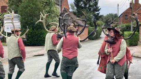 The dancers are dancing in a circle holding the reindeer horns. One who is closest to the camera can be shown holding a carved head of a deer on a wooden pole. 