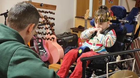 Robin sits in his chair while his dad plays guitar. He is wearing red cord trousers and a striped knitted jumper with buttons down the front. In the background of the photo there is other musical instruments.