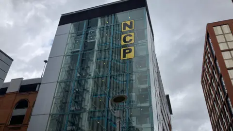 A tall glass building which has a sign that says NCP in black lettering against a yellow background. Behind the building is a cloudy, grey sky