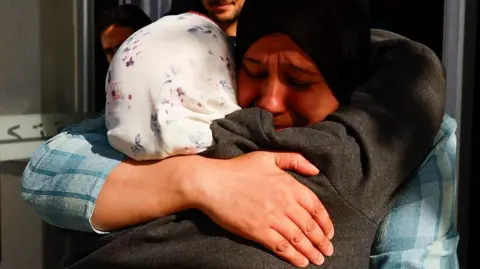 Two women, one with a white patterned headscarf and one with a black one, share an emotional embrace in a doorway. A young bearded man and another person - their face mostly obscured - look on in the background.