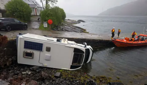 Kyle RNLI A white campervan lying on its side beside a slipway with an orange lifeboat in the right of the picture
