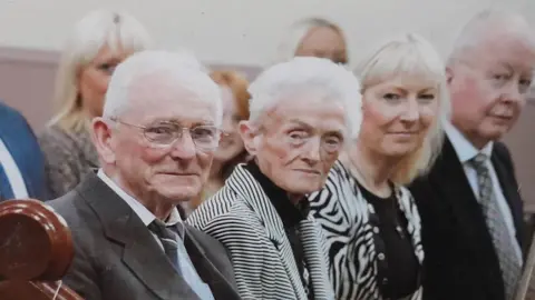 SVLB Billy Short, in a grey suit jacket, white shirt and dark blue tie, is smiling slightly at the camera. His short hair is white and he is wearing glasses. Two women and a man, members of his family, are sitting next to him.