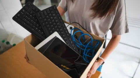 A woman holding a cardboard box of electrical items including two computer keyboards and an iPad. Blue cables can be seen coming out of the box.