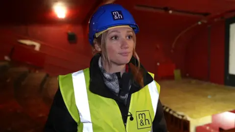 A blond haired woman, wearing a high visibility jacket and blue hard hat, standing in a cinema theatre that is currently undergoing renovation. The walls are red, and the floor brown. She is wearing a black raincoat. 