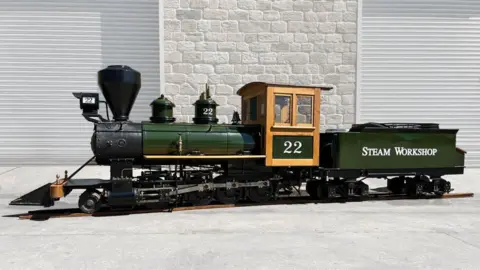 A miniature locomotive in green with Steam Workshop written on it. The number 22 is written on the cab door. Behind the train is a grey brick wall.