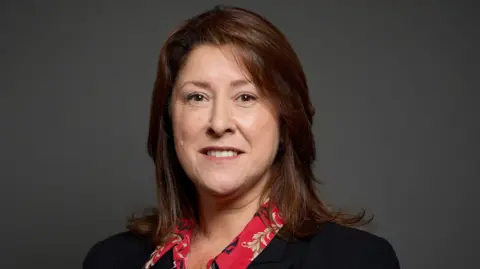 House of Commons/Roger Harris Michelle Scrogham has long brown hair and is wearing a black jacket and red, patterned shirt. She is posing for an official portrait.
