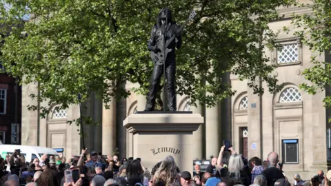 Reuters A crowd, some of them holding up phones, gathers around a beige-coloured plinth which has a black statue on top of Lemmy - a man with long hair holding a bass guitar and singing upwards into a microphone. A large building can be seen in the background.