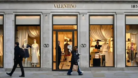 Getty Images People walk past a Valentino store in London. Mannequins dressed in luxury garments are lit-up in its window displays.