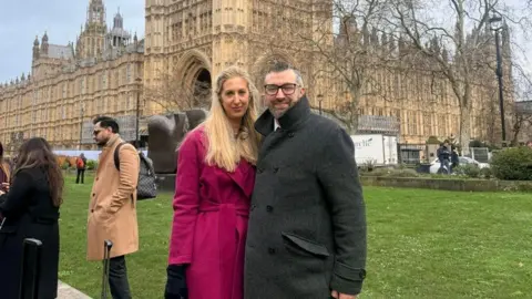 Handout Fiona Winser-Ramm and Dan Ramm stood outside the Houses of Parliament. Mrs Winser-Ramm is wearing a pink overcoat, with Mr Ramm in a grey overcoat.