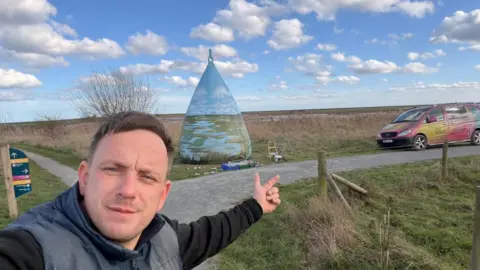 Nathan Murdoch A buoy on grass at a nature reserve. It is painted and features images of a stream. The painting aligns with the horizon. The sky is blue with clouds. A man, wearing a black sweater and a grey bodywarmer is pointing to the sculpture. A pink and yellow van is parked on the grass close to the sculpture. 