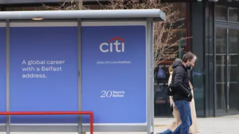 A bus stop with a blue advertisement on it. It says 'A global career with the Belfast address and then the bank Citi's white and red logo. Two people are walking past the bus stop