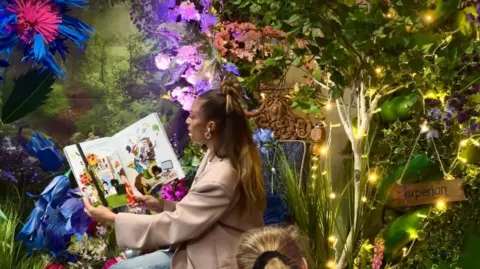 A photo of a woman wearing a blazer and with long brown hair reading a colourful children's book. There is a human made plastic forest behind her.