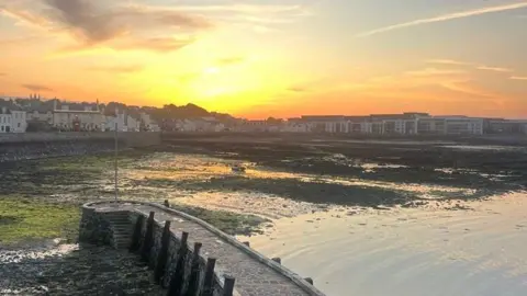 Curved stone path over a seaweed-covered shore at low tide, with waterfront houses and a warm sunset sky in the background.
