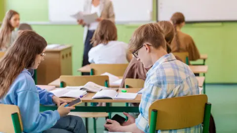 Getty images a teenage boy and a stockshot of a girl were placed in a class with textbooks at a desk and they are looking at their mobile phone. The girl has long brown hair, wearing a blue camber and jeans. The boy has golden brown hair and is wearing black glasses and a blue and white shirt. 