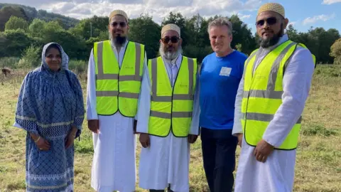 A woman wearing a blue headscarf and outfit alongside four men men wearing florescent vests and one wearing a blue sweatshirt all standing in a grassy field. 