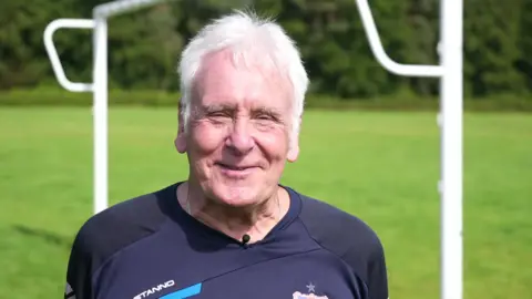 Martin Windle, who has short grey hair. He is stood next to a set of goalposts and is smiling in the photo.