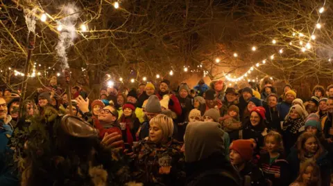 Barley Wood Cider A gathering of people standing under bare trees at night. The trees are lined with pontoon lights and some people are holding fire torches above people's heads.