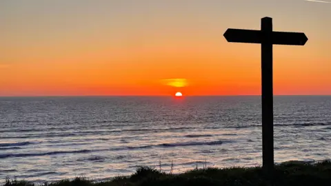 Superted The orange sun popping up over the horizon at the coast. The sky is mostly orange with some blue towards the top. The sea is calm and blue, with some small waves towards the foreground. There is a wooden direction post on the small stretch of land which has been cast in shadow and creates the shape of a cross.